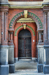 Zabytkowy łuk z czerwonej cegły otacza ciężkie drewniane drzwi z ozdobnymi, żelaznymi moskitierami. Zielone akcenty z płytek, rzeźbione kolumny i zwietrzały kamień tworzą bogatą, ponadczasową fasadę. © Pol Badger