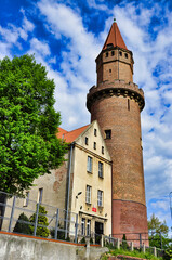 Zabytkowa ceglana wieża i sąsiadujący z nią dom pod czystym, błękitnym niebem, z oświetloną słońcem fasadą i zielonym drzewem - Zamek Piastowski w Legnicy. © Pol Badger