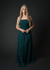 Fototapeta premium Full length portrait of blonde female model wearing royal green ballgown, fantasy crown. Standing, walking pose, front view isolated figure on dark studio background.