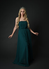 Naklejka premium Full length portrait of blonde female model wearing royal green ballgown, fantasy crown. Standing, walking pose, front view isolated figure on dark studio background.