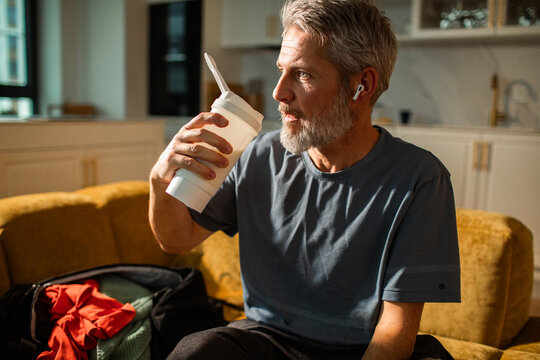 Mature man sipping from shaker cup at home, thoughtful