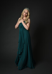 Full length portrait of blonde female model wearing royal green ballgown, fantasy crown. Standing, walking  pose, front view isolated figure on dark studio background.