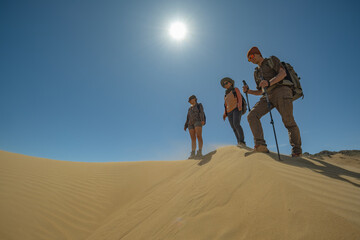 A group of travelers with backpacks and poles stands atop a sand dune under a blazing sun in a cloudless blue sky. A distant low-angle shot captures their isolation.