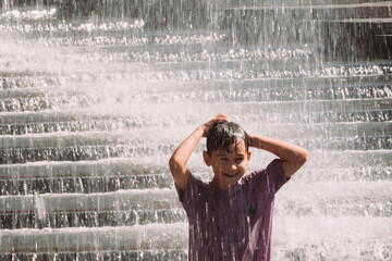 A happy boy in a purple shirt stands under falling water of a fountain or waterfall, enjoying refreshment on a hot summer day