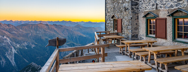 The terrace of Julius Payer House overlooks the stunning Ortler Alps during sunset. Wooden benches line the patio, providing a perfect spot for relaxation and enjoyment of the breathtaking landscape.