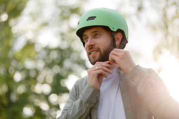 Man adjusts his green helmet while standing outside in a park during a sunny day