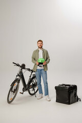 Delivery worker holds helmet while standing next to an electric bike and a delivery bag in a plain studio setting