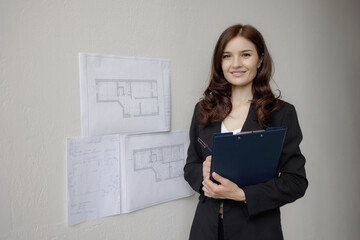 Woman holding clipboard with building plans standing in front of sketches on wall in office during daytime