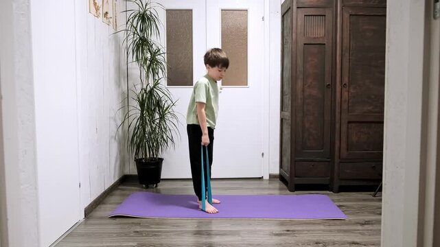 Caucasian boy doing an exercise with an elastic band standing on a yoga mat in a home interior. Pilates workout. Child strengthening the muscles of the back, arms and shoulder girdle. HQ 4k footage.