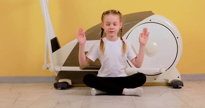 A Caucasian little girl, tired after a workout, sits near an elliptical trainer in a lotus position and meditates with closed eyes against a bright orange wall background. This 4K stock video represen