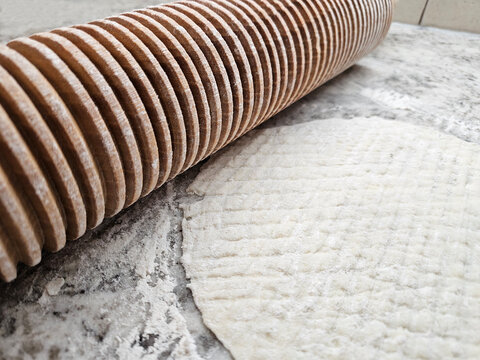 Extreme closeup of a wooden rolling pin with grooves designed specifically to roll out the piece of lefse flatbread that is on the kitchen countertop next to the rolling pin.