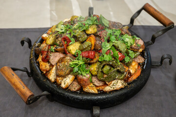 A large, sizzling sach platter filled with roasted meats, potatoes, bell peppers, sun-dried tomatoes, and fresh herbs, presented on a rustic cast iron pan.