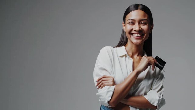 A young woman smiling while holding a credit card, standing against a plain background, casual pose with one arm crossed and the other hand holding the card up, confident and cheerful expression