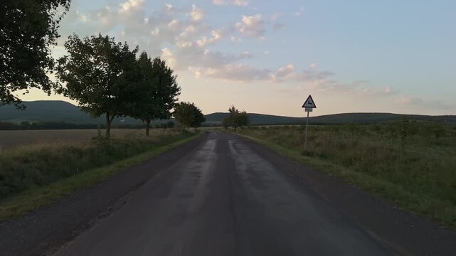 Low-level fly on a bumpy rural road leading to Vilyvit&aacute;ny, Hungary, Zempl&eacute;n region at sunset, bordered by trees and fields with a deer-crossing warning sign adding context to the tranquil countryside.