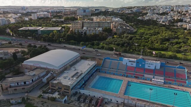 Malta national swimming pool, university, and Mater Dei Hospital buildings from a drone perspective, split by a busy road surrounded by a green park, on a sunny winter day in Msida.