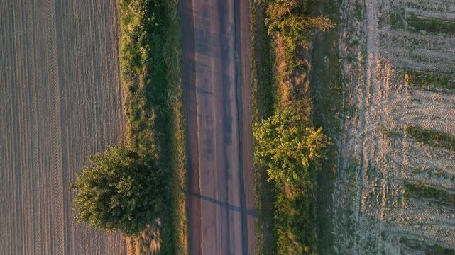 Tilt-down drone view of a bumpy rural road winding through agricultural fields and trees in Zempl&eacute;n, Hungary, leading to Vilyvit&aacute;ny, captured during the golden hour of an autumn sunset.