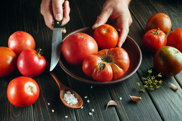 Hands are cutting ripe tomatoes on a wooden board. A bowl holds more tomatoes, and salt and garlic are nearby. This scene shows a kitchen setting