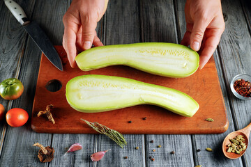 A person holds two pieces of sliced cucumber on a wooden board. A knife rests nearby while tomatoes, herbs, and spices are arranged around the board. The scene is set in a kitchen