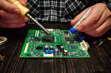 A person uses a soldering iron and other tools to repair a circuit board on a wooden table. The setting is a workshop with dim lighting and various tools nearby © Віталій Б.