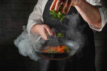 A chef adds fresh herbs to a pan of cooking carrots. Steam rises from the pan as the chef focuses on the task. The kitchen has a rustic brick wall backdrop