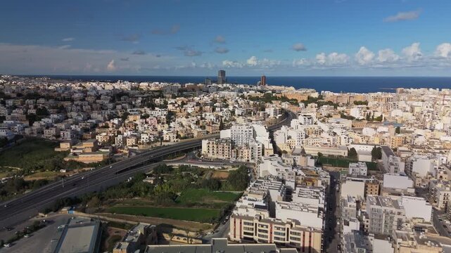 Forwarding drone view from Msida, Malta, on a sunny winter day with moving cars and clouds over the bustling skyline of Kappara, San Ġwann, St Julian's and Swieqi neighborhoods.