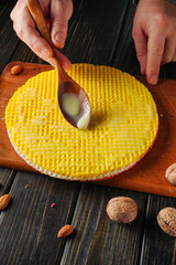 The photograph shows a hand spooning condensed milk onto a round waffle piece lying on a wooden board. Nuts are scattered on the board. Cake making is taking place in the kitchen.
