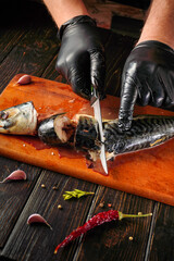 A person cuts a fish mackerel with a knife on a wooden surface. Garlic, pepper, and chili are present nearby. The hands are gloved for hygiene