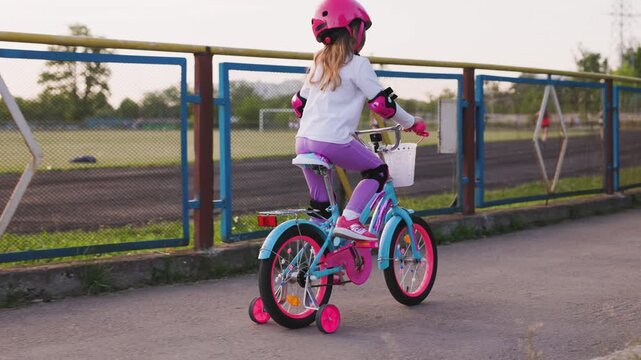 Little girl riding a small bicycle with training wheels, wearing helmet and protective gear, cycling along a sports track on a warm summer day