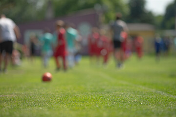 low angle grass with orange ball in foreground and blurred youth teams in background, wide open pitch during community soccer session, warm