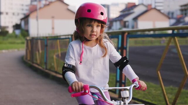 Little girl confidently riding a children bicycle with helmet and protective pads on an urban park path, learning balance and safe cycling in a residential area