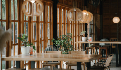 A cozy wooden caf&eacute; interior filled with warm natural light, featuring minimalist furniture, hanging pendant lamps, large windows, and green plants that create a calm and inviting atmosphere.