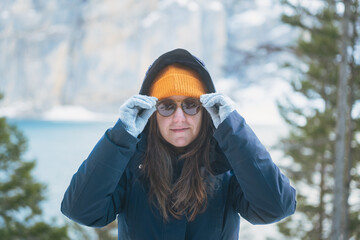 Front view of a young woman in a hooded blue parka adjusting her sunglasses with grey gloves on a winter day