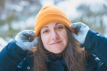 Frontal portrait of a smiling woman adjusting her orange winter beanie with grey wool gloves