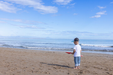 Little boy playing with a big red shovel on a sandy beach near sea. Summer holidays concept.