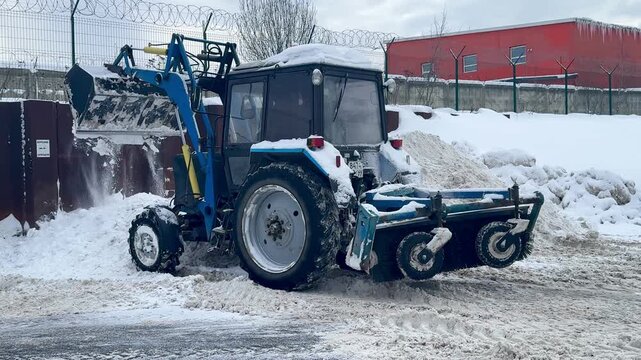 A blue MTZ (Belarus) tractor with a front loader clears snow at a large delivery warehouse on a snowy winter day, showing industrial maintenance operations.