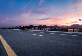 Empty asphalt road and modern city skyline with mountain range under a dramatic sunset sky. © zhao dongfang