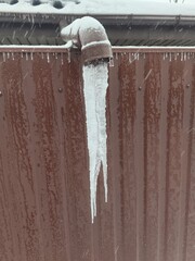 Large sharp icicle hanging from a gutter pipe on a brown fence background