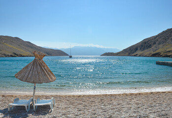 A scenic wide view of a straw sun umbrella and white lounge chairs on a pebble beach at Vela Luka on Krk island overlooking a bright blue bay
