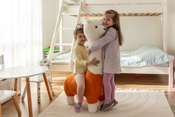 Cheerful little sisters playing together at home, hugging a large plush llama toy in their cozy kids' room. Happy childhood and family bonding concept.