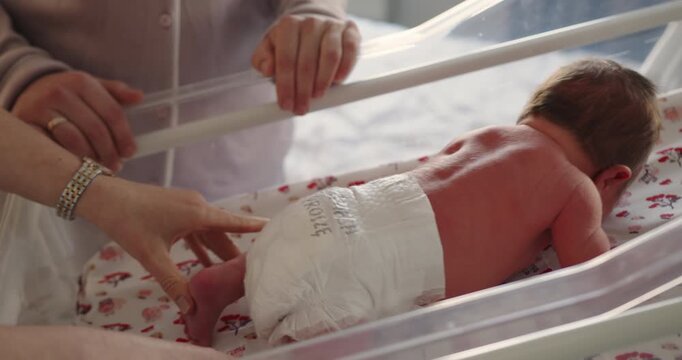 Newborn infant rests in clear plastic bassinet with parents' hands nearby showing care and tenderness