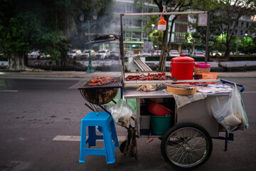Grilling various meat for customers on mobile stall with homemade grill outdoors at street food market in Bangkok. Cooking and selling unhealthy snacks, junk food and local cuisine of Thailand. © DimaBerlin