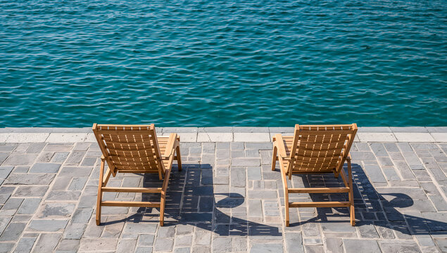 Two empty wooden lounge chairs facing the calm blue ocean on a sunny day - Powered by Adobe