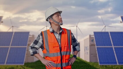 Asian Male Engineer Wearing Safety Helmet Looking Around While Standing With Arms Akimbo at Energy Farm featuring Solar Panels, Battery Storage and Wind Turbines