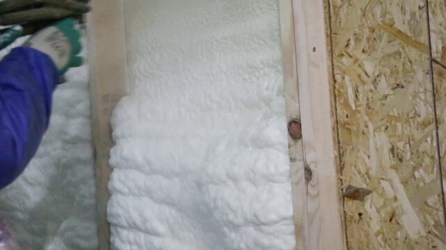 A construction worker in overalls applies insulation foam to a wooden wall of a house, house construction and insulation
