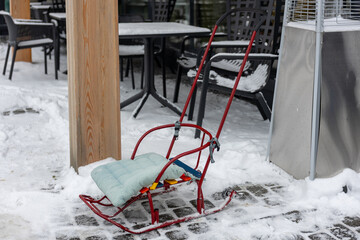Red metal sled with cushion seat standing on snowy pavement near outdoor cafe in winter