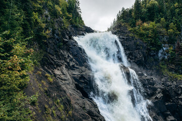 Obraz premium Ouiatchouan Falls cascading through green forest with observation deck at Val-Jalbert Historical Village in Quebec