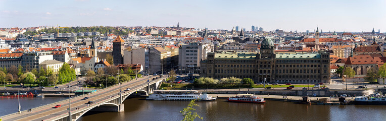 Obraz premium Sweeping panoramic view of Prague across the Vltava River, showcasing a historic bridge, red rooftops, and grand buildings along the riverfront. Ideal for travel, cityscape, and architectural themes.