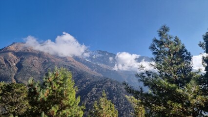 Majestic mountain peak shrouded in swirling clouds against a vibrant blue sky, with lush green pine...