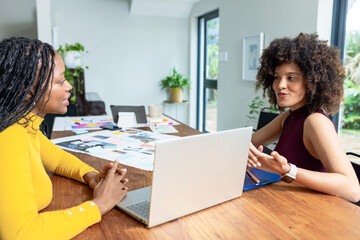 Diverse female coworkers sitting at home, discussing printed layouts with laptop and coffee mug