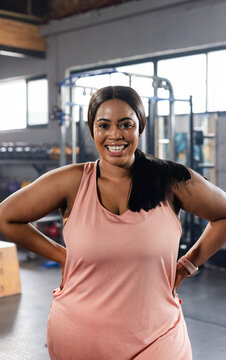 African American mid-30s woman posing hands-on-hips smiling in gym with squat rack pink tank watch